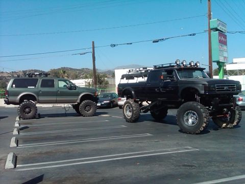 1986 Ford Bronco - A heavily modified black Ford F-Series truck, likely from the early 1990s, is parked next to a lifted green SUV.