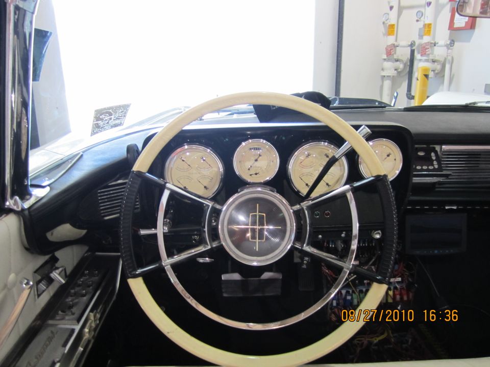 Interior shot of a vintage car dashboard with a large steering wheel and multiple gauges. The center of the steering wheel has a logo