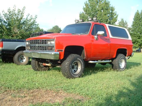 1994 GMC Suburban - A red SUV with a white top and off-road tires sits on a grassy field next to another truck.