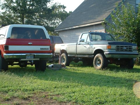 1994 GMC Suburban - Two trucks, a Ford F-Series and a Chevy K5 Blazer, sit parked in a yard on a sunny day.