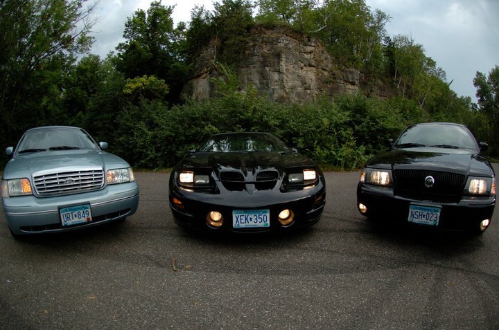Light blue Ford Crown Victoria, black Pontiac Firebird, and black Mercury Grand Marquis parked in a row