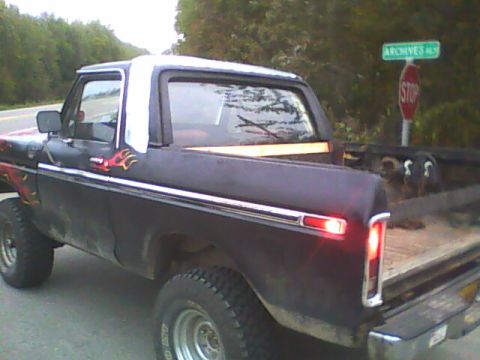 1978 Ford Bronco - Weathered dark blue/black lifted Ford 4x4 with a white half-cab top and open bed, shot from the left rear three-quarter at a roadside stop…
