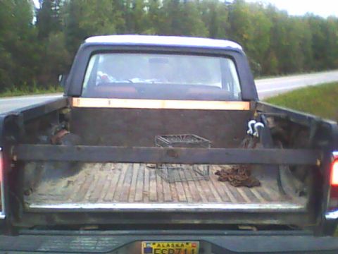 1978 Ford Bronco - Rear view of a dark early Ford Bronco half-cab with the tailgate lowered, revealing a weathered wood-planked bed holding a wire basket and…