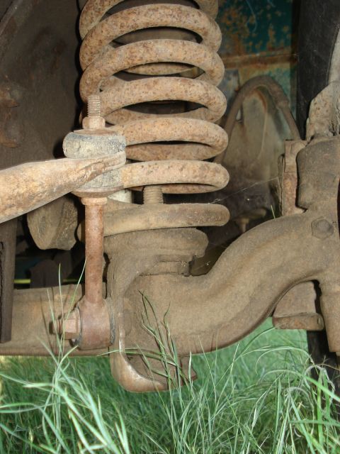1979 Ford F-150 - Close-up of a heavily rusted front suspension coil spring, sway-bar link and steering arm above grass.