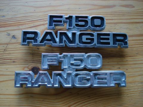 1979 Ford F-150 - Two sets of chrome and black Ford F-150 Ranger emblems are displayed on a wooden background.