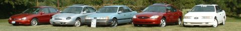 1993 Ford Taurus - Five mid-size sedans lined up on grass at a car show, including red, silver, blue, maroon and white examples.