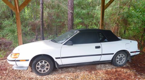 1988 Toyota Celica - A white 1988 Toyota Celica convertible is parked outdoors under a wooden structure.