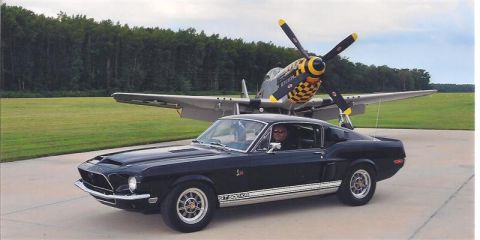 1968 Shelby GT500 - A black 1968 Shelby GT500 is parked on an airfield with a P-51 Mustang airplane behind it.