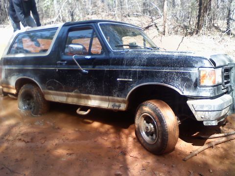 1990 Ford Bronco - Muddy 1990 Ford Bronco is stuck off-road, with a person attempting to push it free.