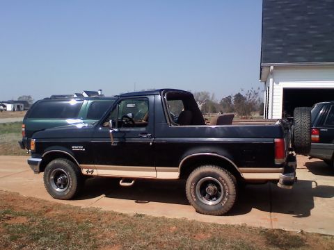 1990 Ford Bronco - This black 1990 Ford Bronco features tan two-tone trim and a rear-mounted spare tire, parked next to other vehicles.