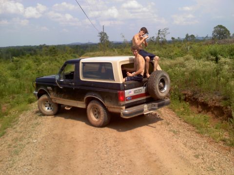 1990 Ford Bronco - Two young men ride in the back of a 1990 Ford Bronco on a dirt path with green foliage around.