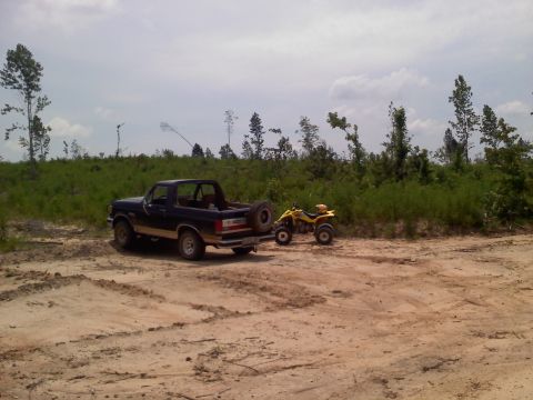 1990 Ford Bronco - Ford Bronco and ATV in a dirt lot with trees and brush in the background.