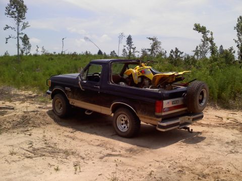 1990 Ford Bronco - A Ford Bronco is parked on a dirt path, carrying a yellow ATV in its open bed.