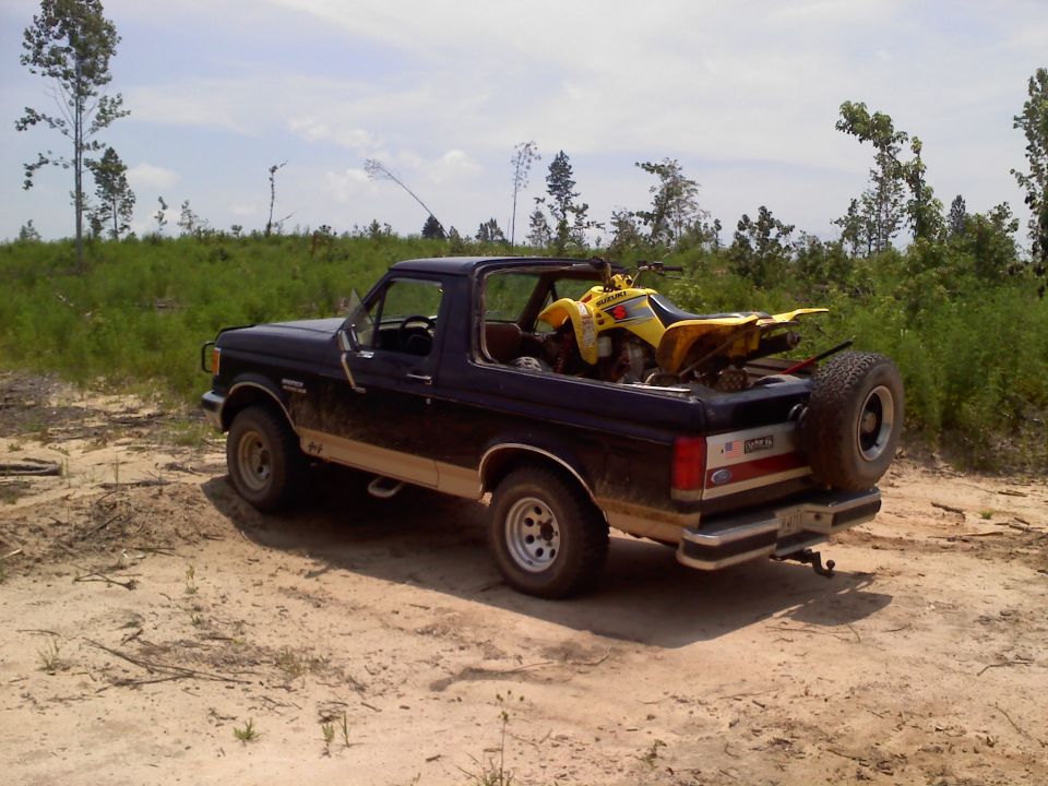 Dark blue Ford Bronco parked on dirt road, yellow ATV in the bed. Trees and brush in background