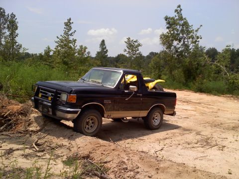 1990 Ford Bronco - A black Ford Bronco from the late 1980s is parked on a dirt path with a yellow ATV loaded in its bed.