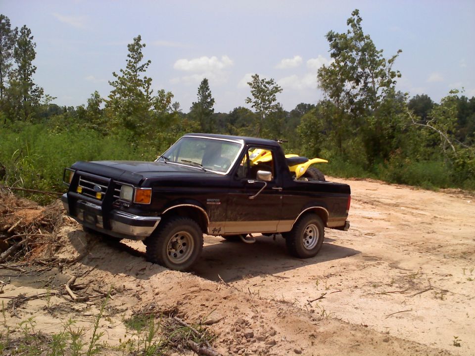 Black Ford Bronco, 1987 model year, with a brush guard and a yellow ATV in the bed, angled from the front-left on a dirt road