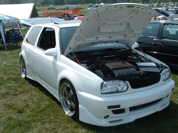 White hatchback with hood open, custom body kit, aftermarket wheels, and visible engine