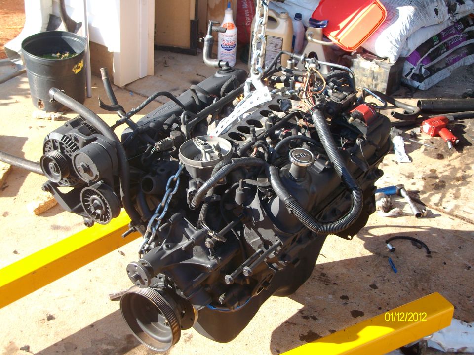 Engine assembly on yellow engine hoist, with various hoses, wires, and a pulley system visible