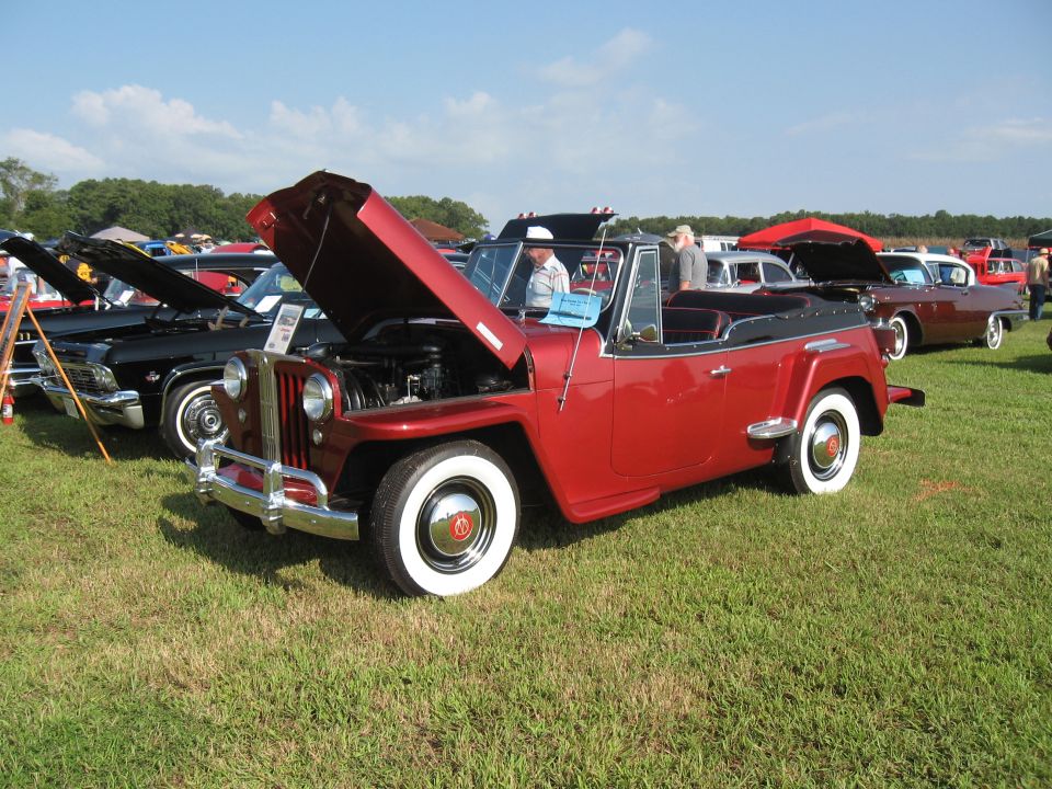 Red 1955 Jeep CJ-5 convertible with hood open, parked on grass at a car show with other vehicles