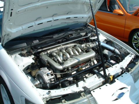 1993 Ford Taurus - The engine bay of a white car is open, revealing a V6 engine with a turbocharger. An orange car is visible in the background.