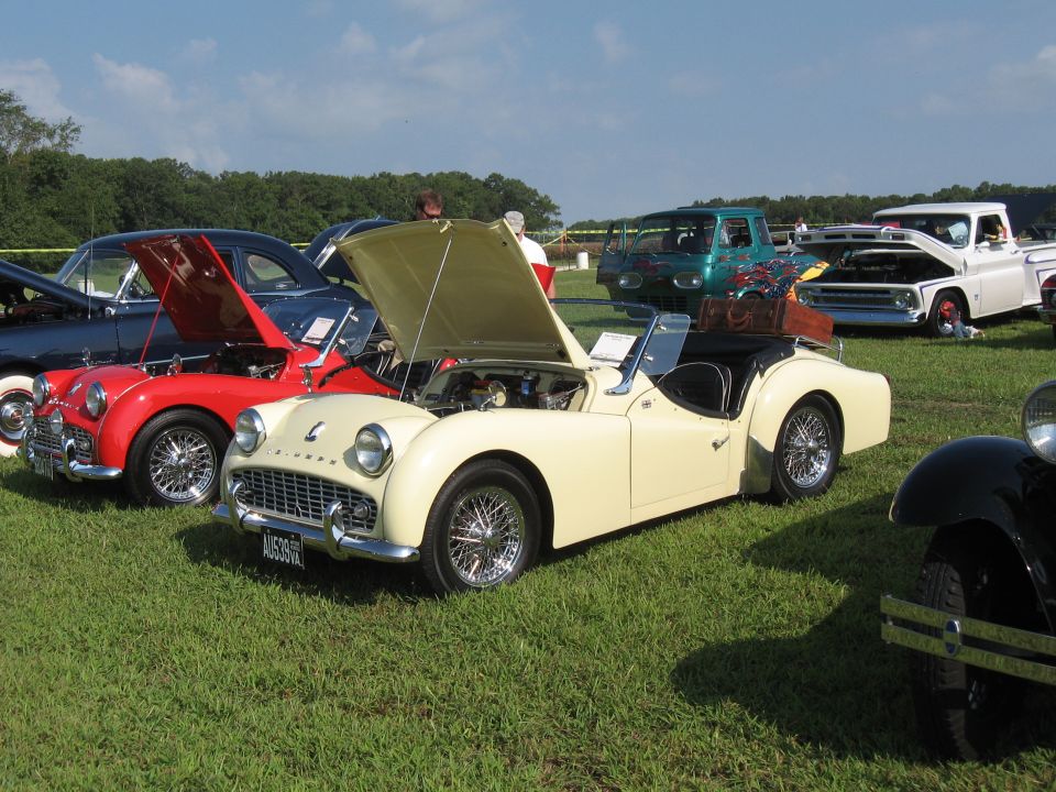 Cream-colored Triumph TR3 with hood open, parked on grass next to a red Triumph TR3. Other cars and trucks in background