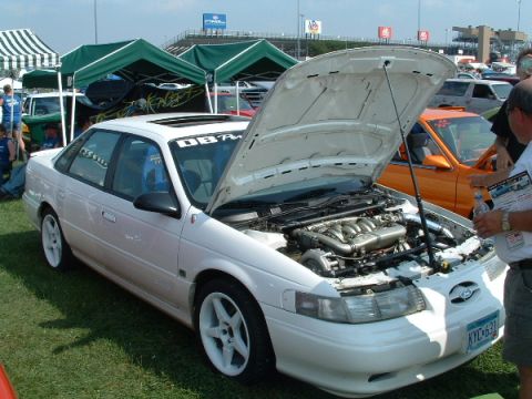 1993 Ford Taurus - A white sedan is displayed with its hood open at an outdoor event, revealing its engine and turbocharger.