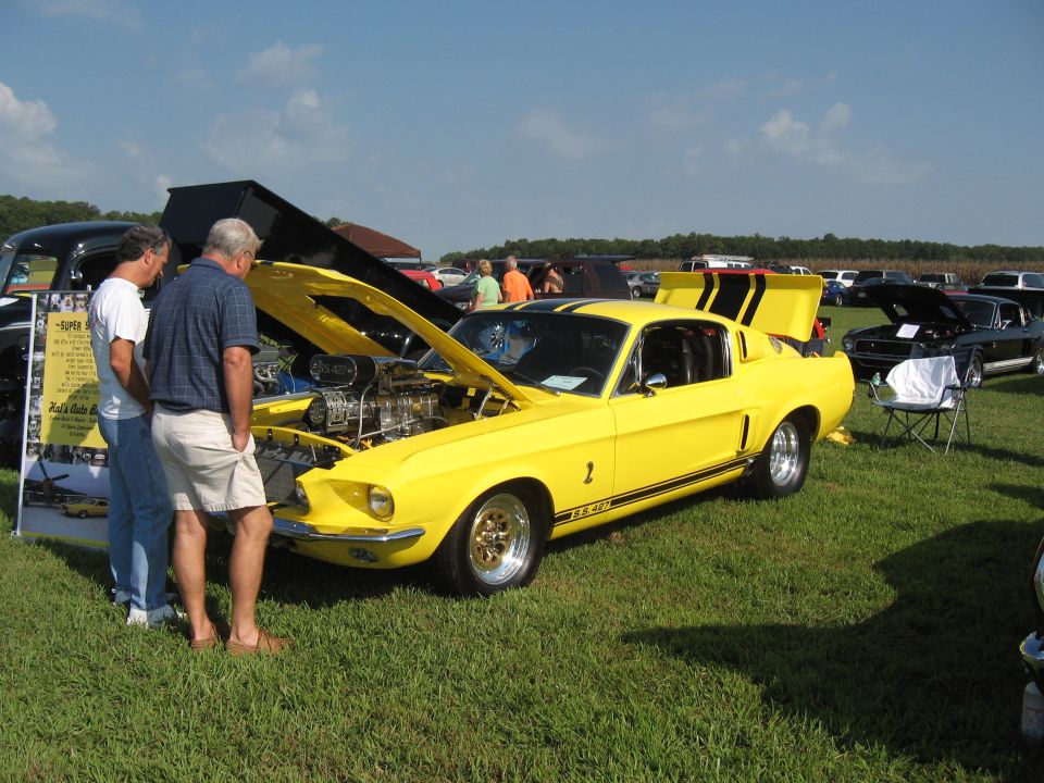 Yellow 1968 Shelby GT500 with hood and trunk open, two men looking at engine. Other cars in background