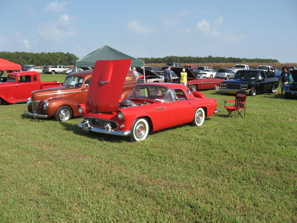 Red 1957 Ford Thunderbird convertible with hood open, parked on grass at a car show