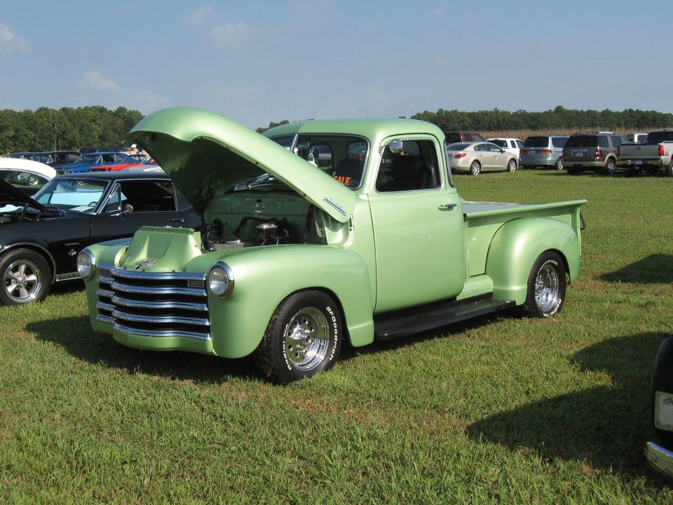 Light green pickup truck with hood open, parked on grass at an outdoor event. Another car is visible to the left