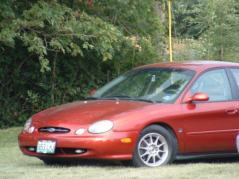 1993 Ford Taurus - Red Ford Taurus parked on grass with trees in background.