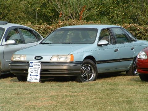 1993 Ford Taurus - A light blue sedan is parked on grass next to another vehicle, with a sign indicating reserved parking for a Taurus Car Club.