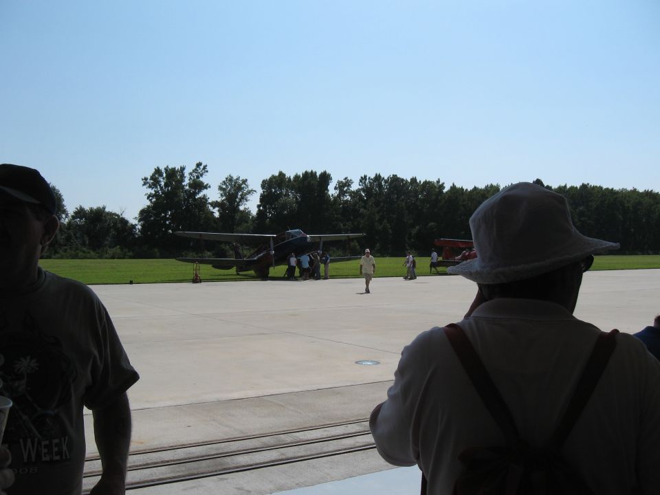 Airplane on tarmac with people around it, viewed from behind a person wearing a hat
