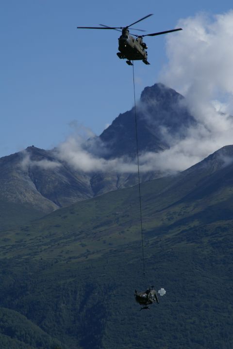 1990 Chevrolet Suburban - A military helicopter transports a smaller vehicle suspended by a cable, with a mountainous, forested backdrop under a blue sky.