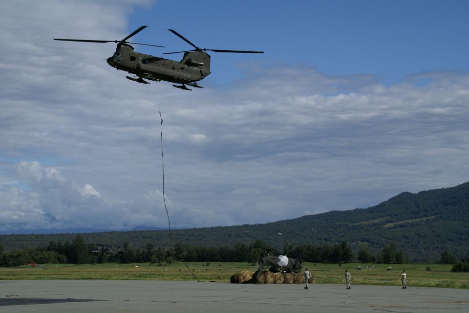 Military helicopter in flight, suspended load below, ground crew visible near cargo
