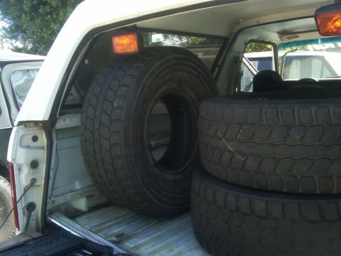 1986 Ford Bronco - Multiple spare tires are visible in the bed of a truck, with another vehicle partially seen in the background.
