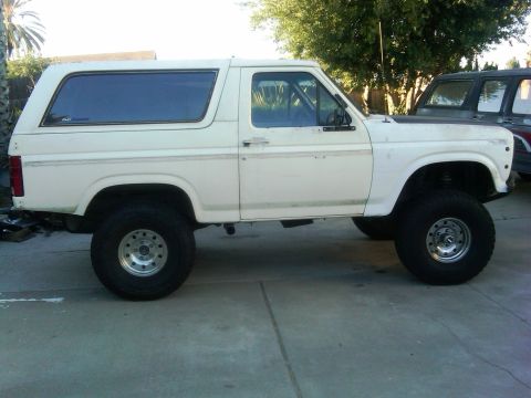1986 Ford Bronco - This white SUV, likely from the early 1980s, is shown from the side with large off-road tires and aftermarket wheels. Another vehicle is pa…