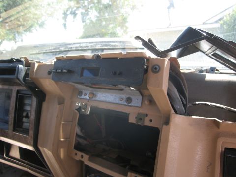 1986 Ford Bronco - Close-up of a tan 1980s Ford truck dash with the radio and trim removed, exposing the open center stack and wiring while a black digital cl…