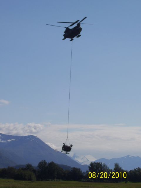 1990 Chevrolet Suburban - A helicopter transports a chair via cable over a landscape with mountains and trees.