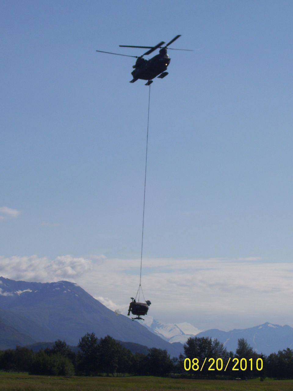 Helicopter carrying a chair suspended by a cable over a grassy field with mountains in the background
