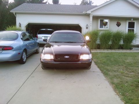 2003 Ford Crown Victoria - Front view of a dark sedan, possibly a 2003 Ford Crown Victoria, parked in a driveway with a spotlight on the driver's side mirror.