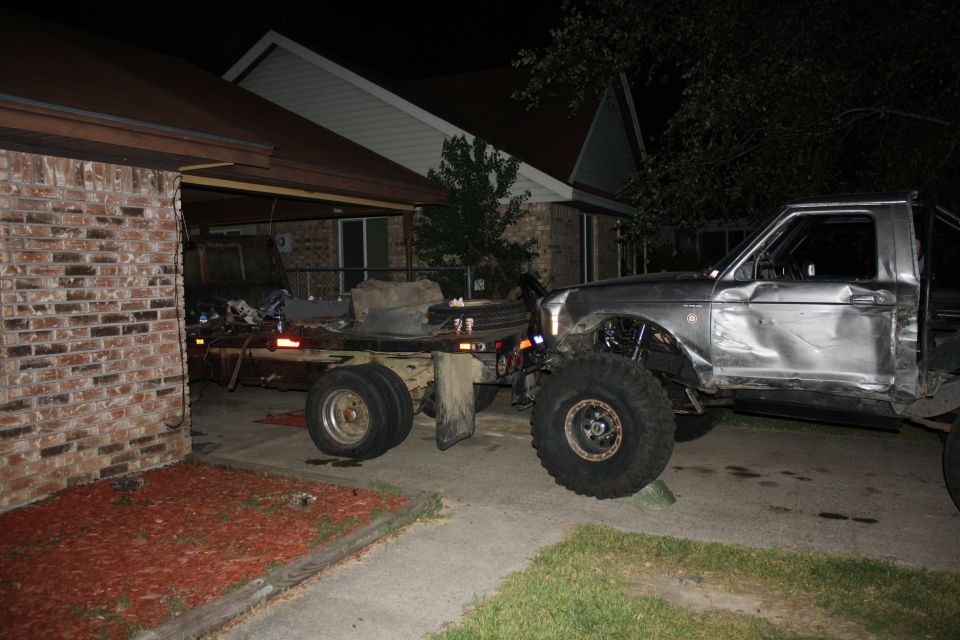 Damaged silver pickup truck with large tires next to a flatbed trailer parked in a driveway at night