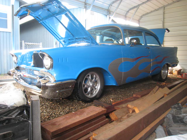 Light blue tri-five Chevy sedan with ghost flames and billet wheels, hood up in carport