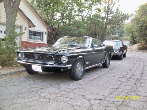 1968 Ford Mustang - A dark green 1968 Ford Mustang convertible is parked on a cracked road, with another car behind it.