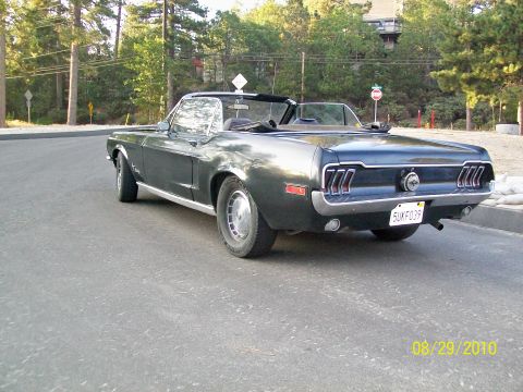 Dark green Ford Mustang convertible, top down, rear 3/4 view on tree-lined street.