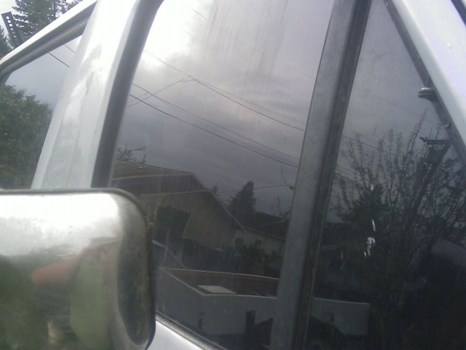 Side view mirror and tinted window of a vehicle, reflecting houses and trees