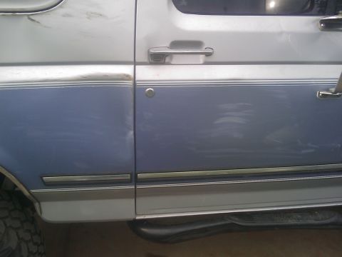 1996 Ford Bronco - Side view of a damaged two-tone blue and silver Ford Bronco, revealing scratches and dents on the door panel.