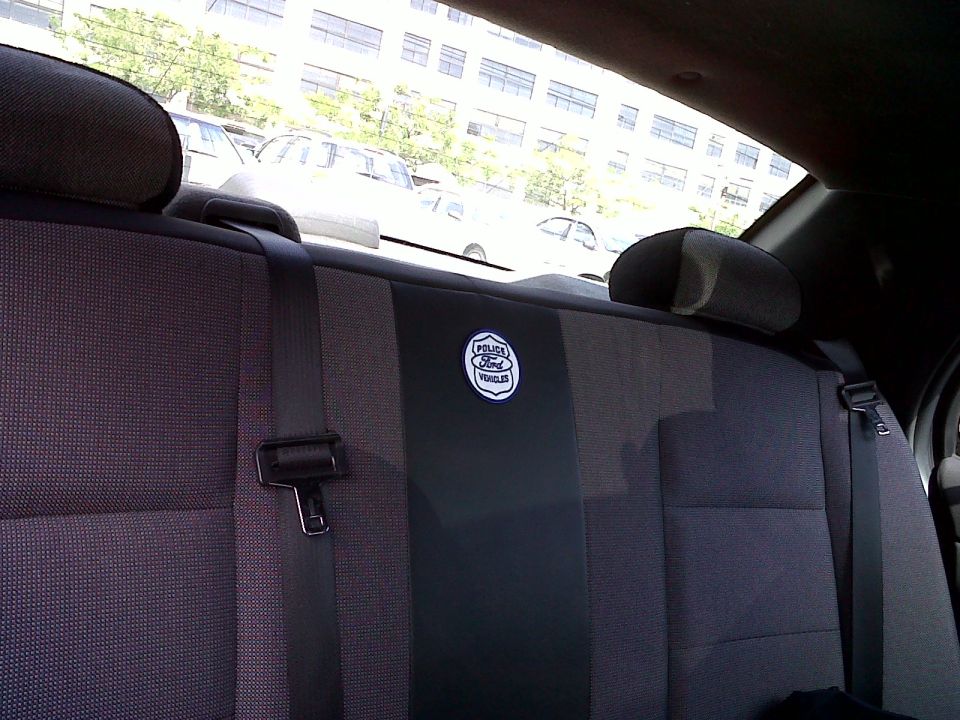 Rear seat interior of a car, showing a Ford Police Vehicles badge on the seatback. Other vehicles are visible through the window