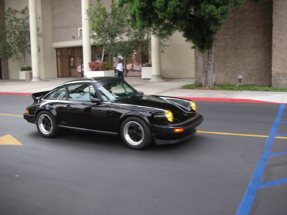 Black Porsche 911 with a whale tail spoiler, driving on asphalt with yellow and blue markings