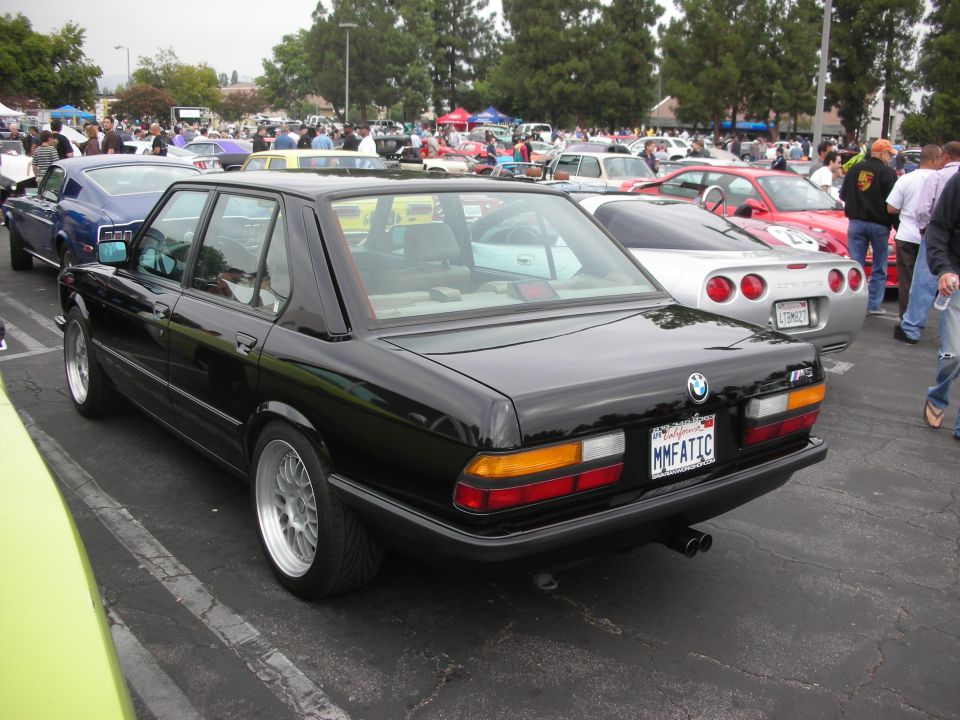 Black BMW E28 sedan with M5 badge, parked at a car meet. Silver Corvette visible behind