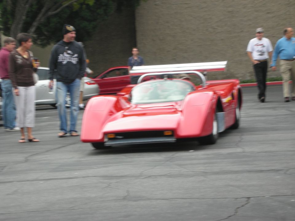 Red race car with a large rear wing, parked on asphalt with people around